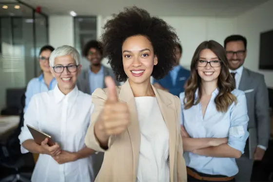 A group of men and women smiling in office setting and woman in front giving thumbs up.