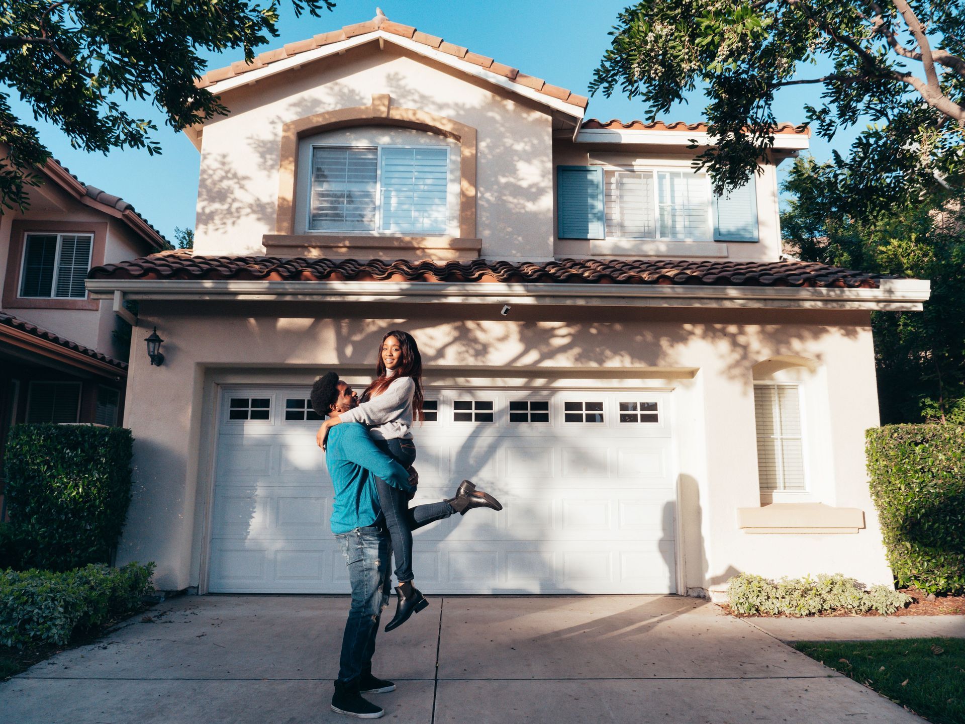 Person lifting another in front of a two-story house with a white garage door