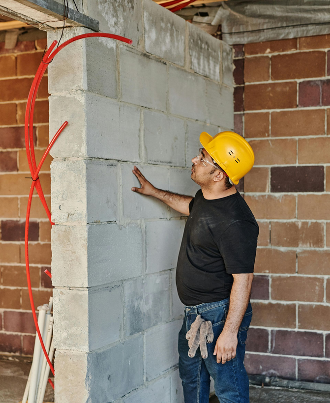 Corner of a residential building showing foundation damage with cracked concrete and visible underlying brickwork.