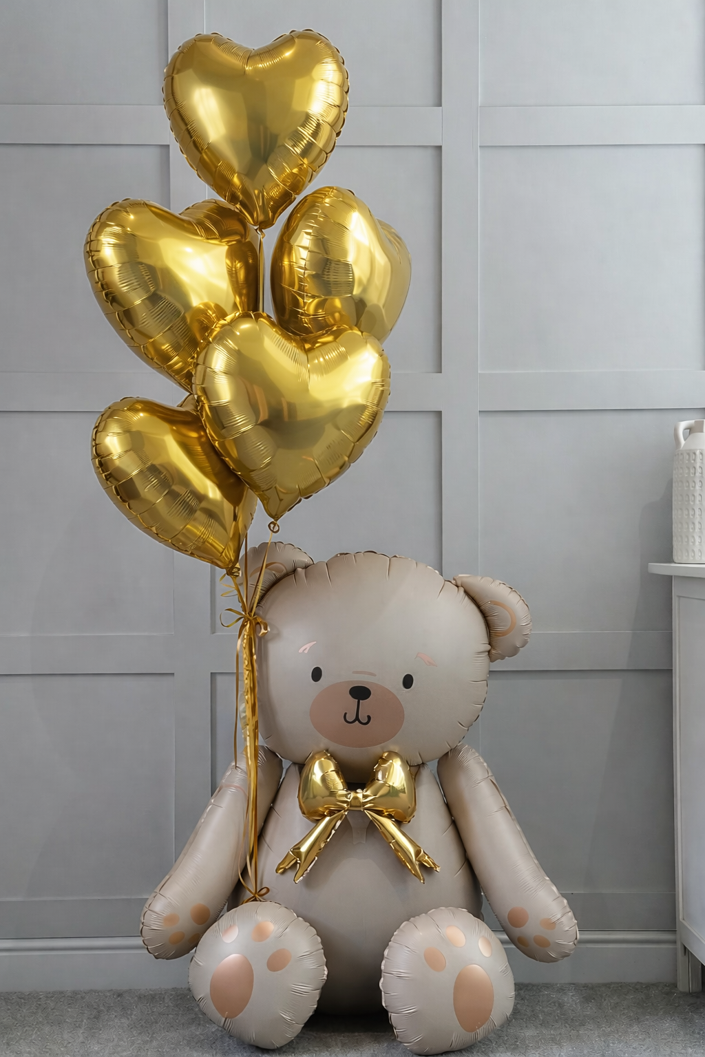 A beige teddy bear balloon sitting on the floor, holding a bunch of five shiny gold heart-shaped balloons.