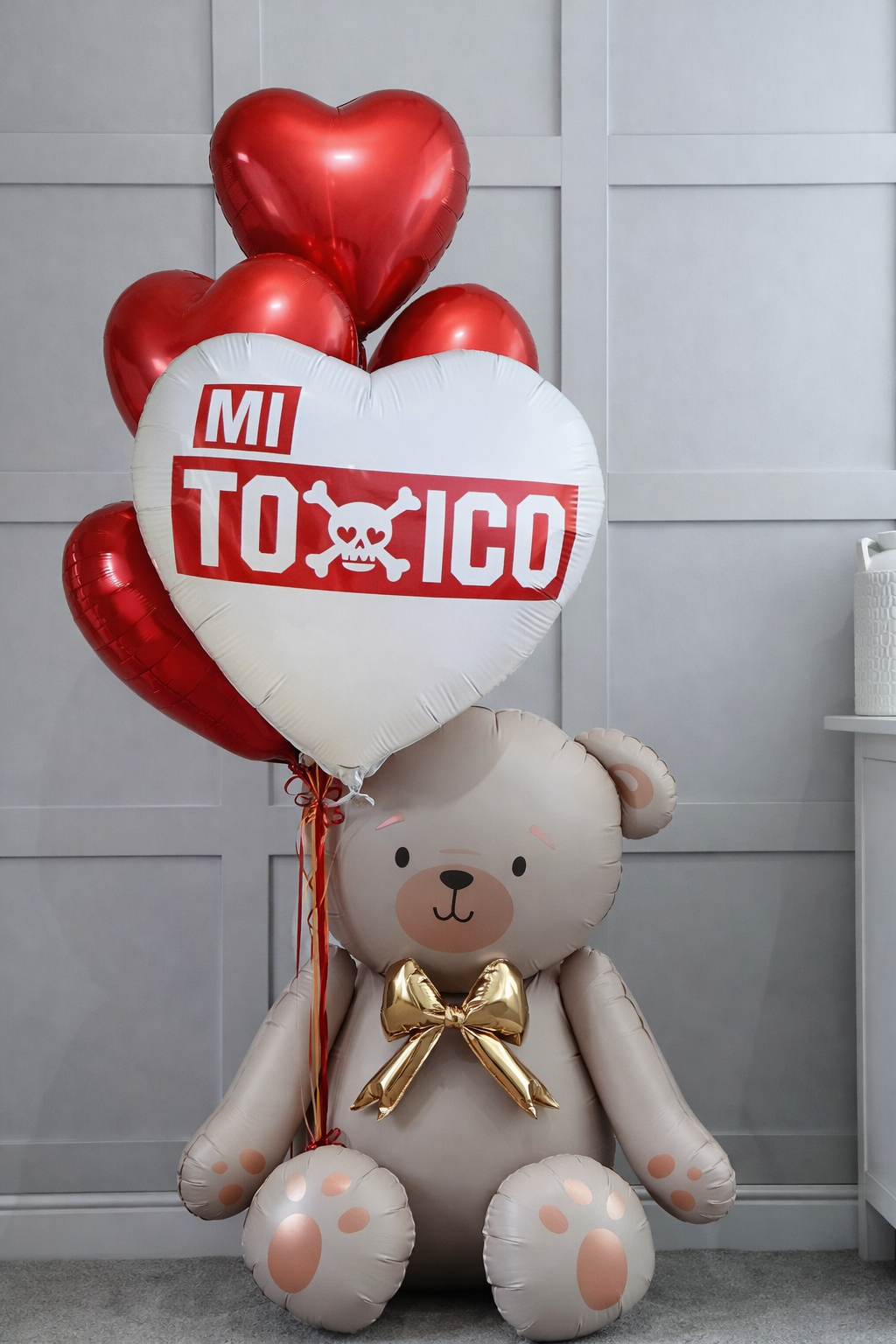 A light brown teddy bear balloon sits holding a bouquet of red heart-shaped balloons, one with the text