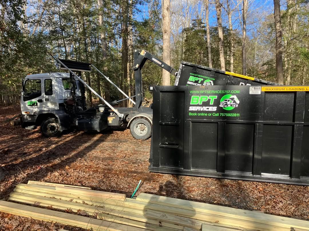 Truck loading a black dumpster in a wooded area. The truck has a crane-like arm and the dumpster has the logo 