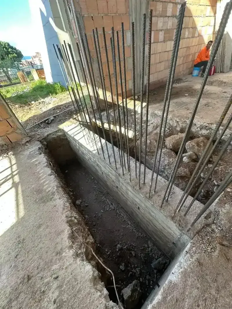 Cantiere con trincea in cemento e barre di armatura che si estendono verso l'alto, vicino a un edificio in mattoni.