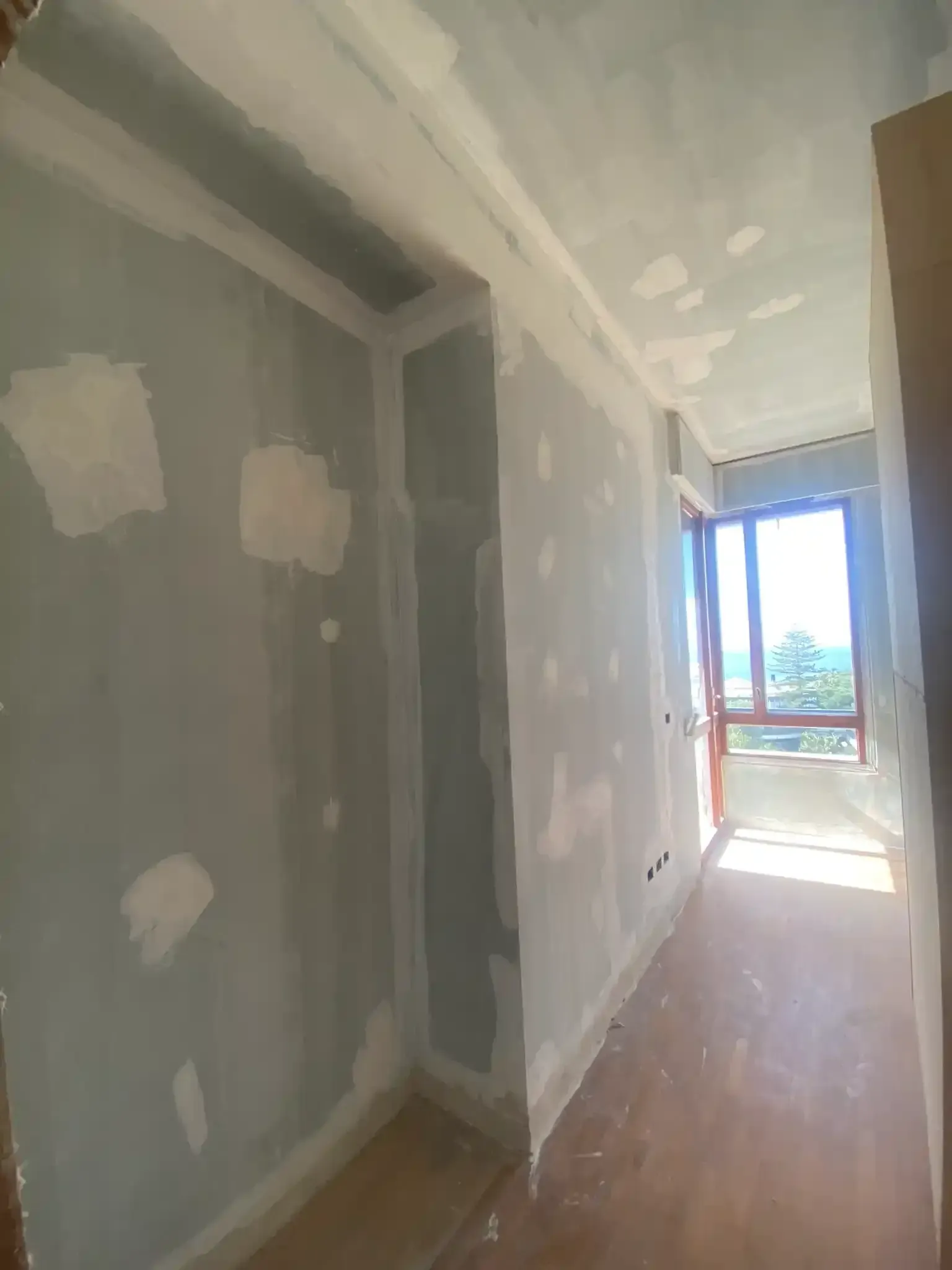Corridoio interno in costruzione; cartongesso, stucco e pavimento in legno grezzo. Luce solare che filtra da una finestra.