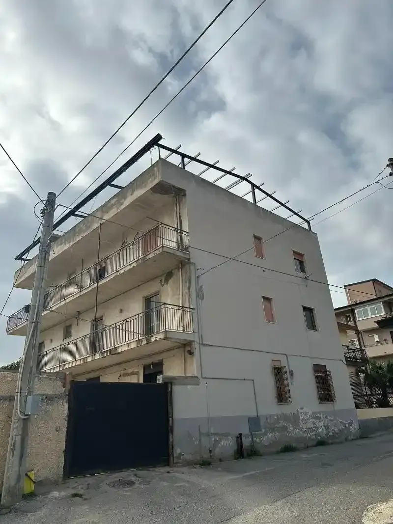 Un edificio di tre piani con balconi, facciata grigia e linee elettriche aeree. Davanti c'è un grande cancello nero.