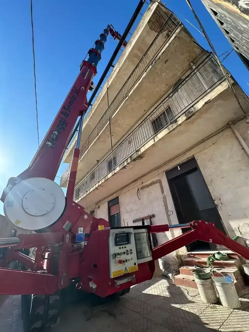 Una gru rossa si protende verso un edificio di tre piani, cielo azzurro, parcheggiato sulla strada.