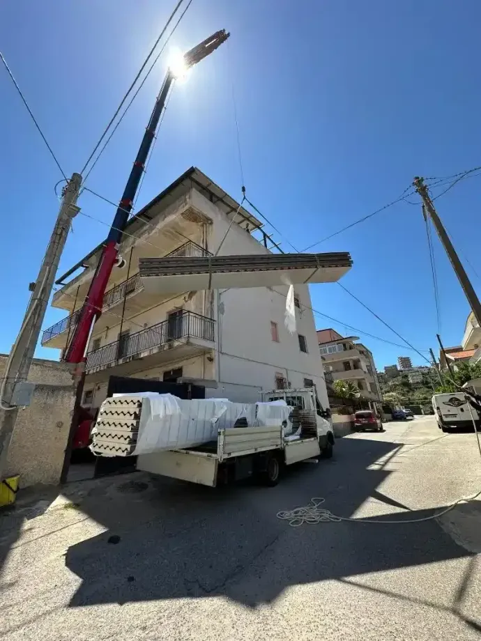 Una gru solleva una lastra di cemento da un edificio di tre piani, passando sopra un camion su una strada soleggiata.