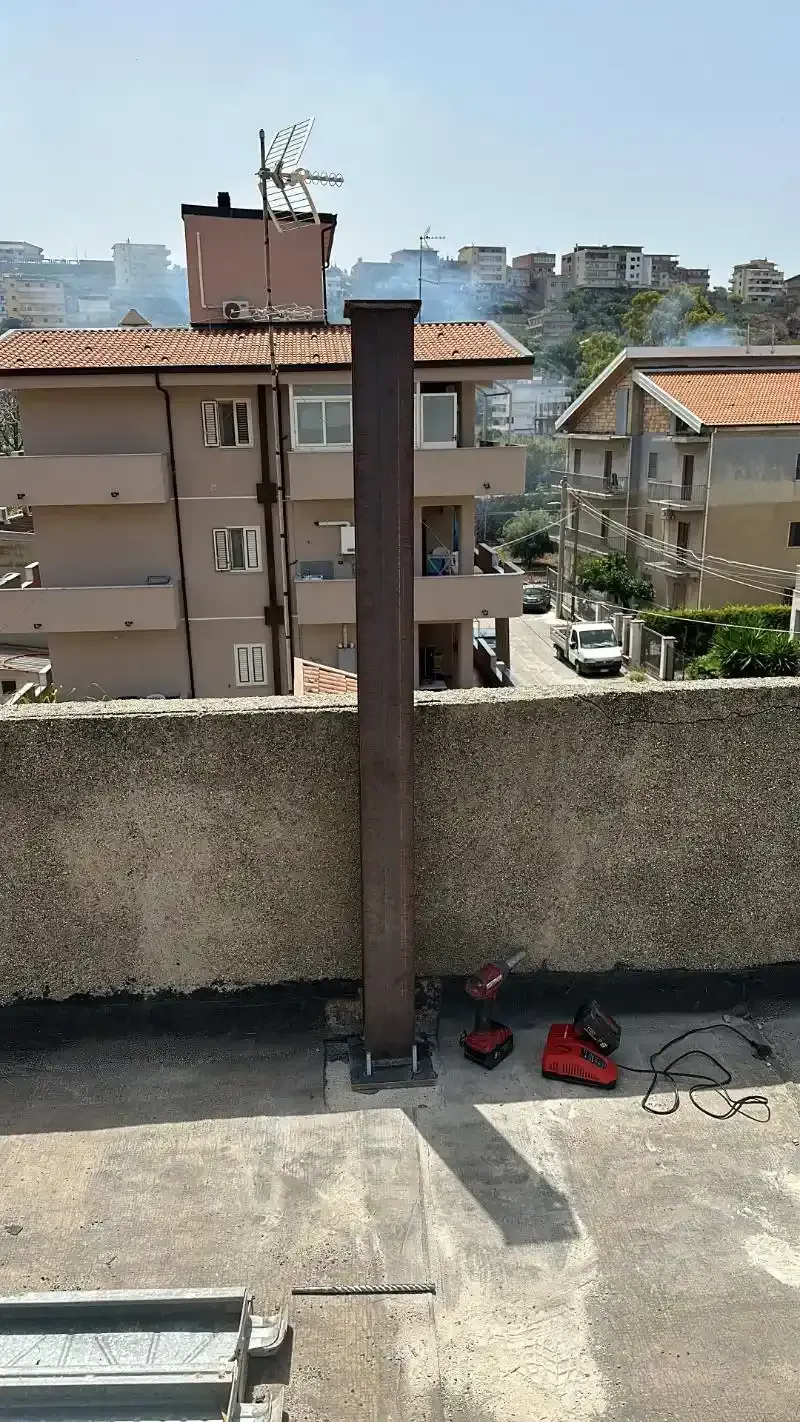 Un alto camino metallico si erge su un tetto, con edifici cittadini e cielo azzurro sullo sfondo.