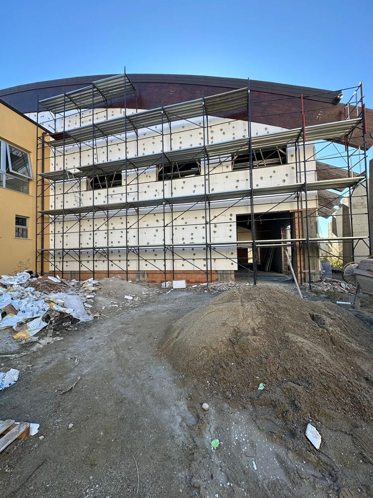 Edificio in costruzione; muri beige, impalcature, reti verdi, cielo azzurro.