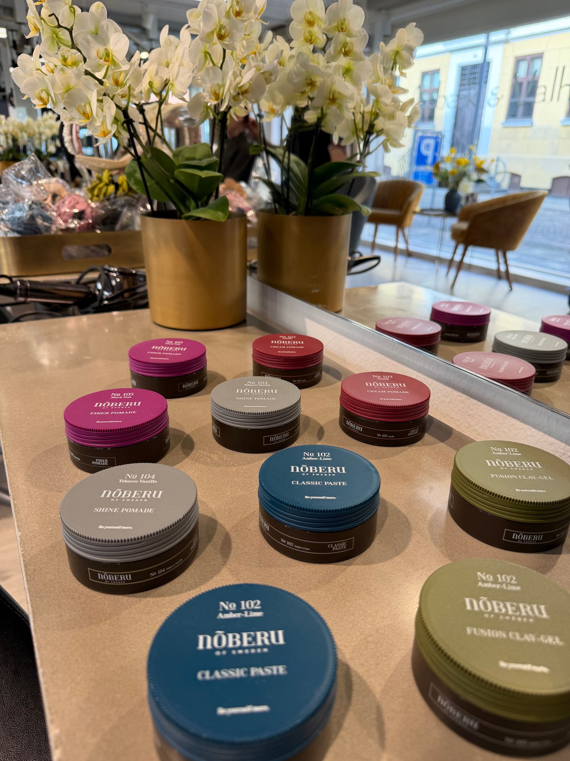 Udstilling af runde hudplejekrukker og -dåser på en disk ved siden af ​​hvide blomster og et spejl i en lys butik.