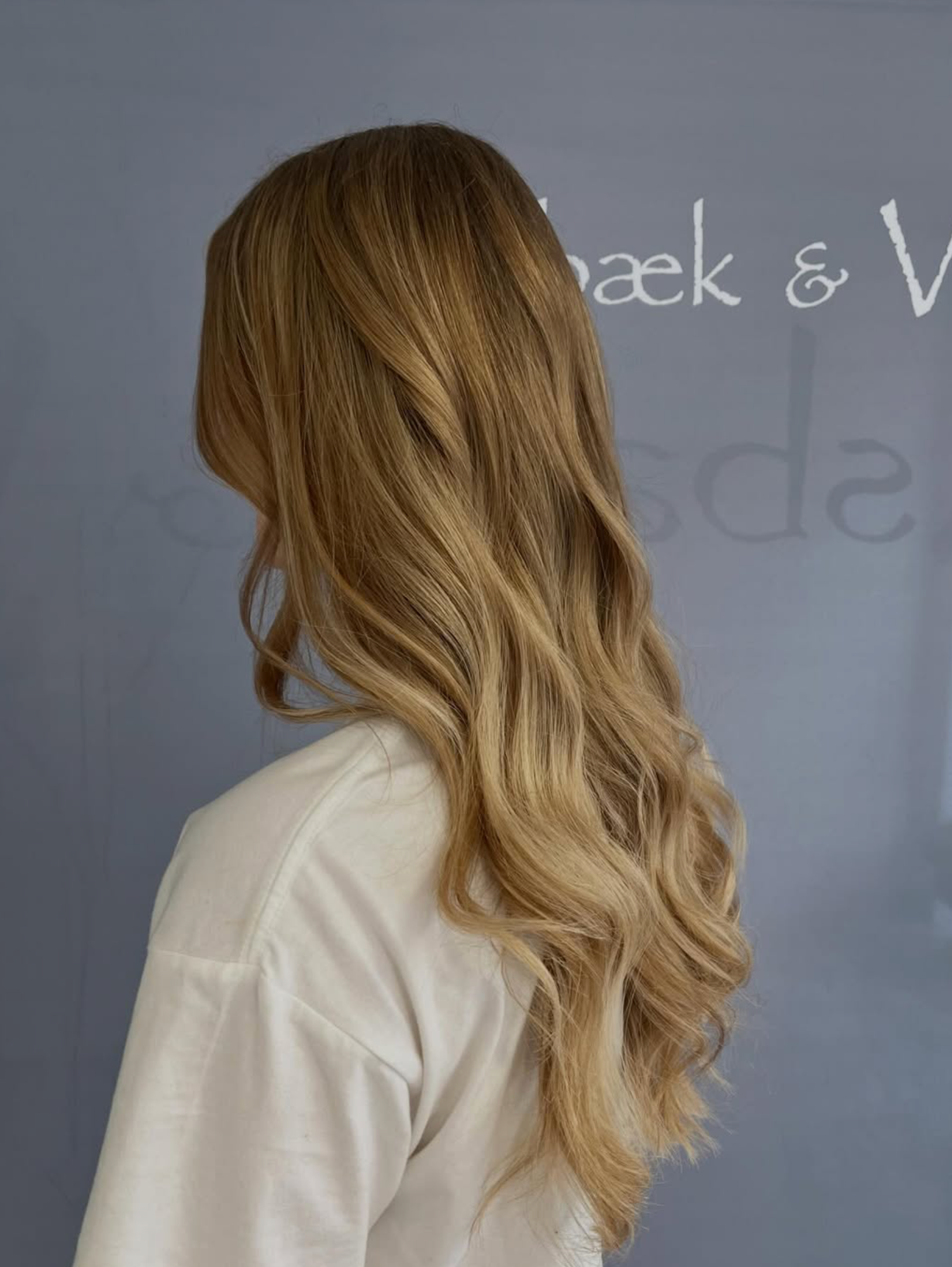 Bagfra set langt, bølget blond hår på en person i en hvid skjorte mod en grå baggrund
