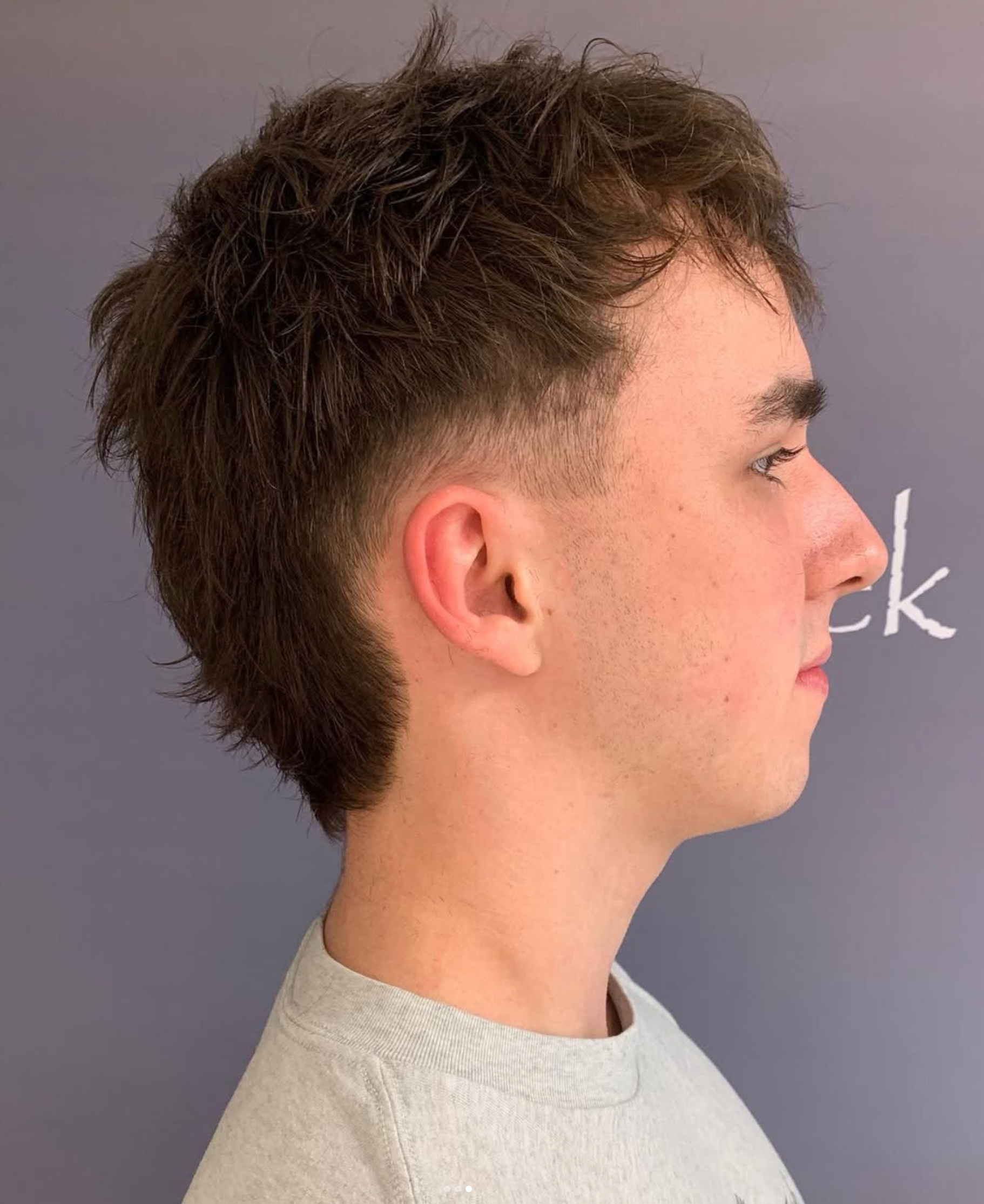 Sideprofil af en person med kort brunt hår iført en lysegrå skjorte mod en grå baggrund.
