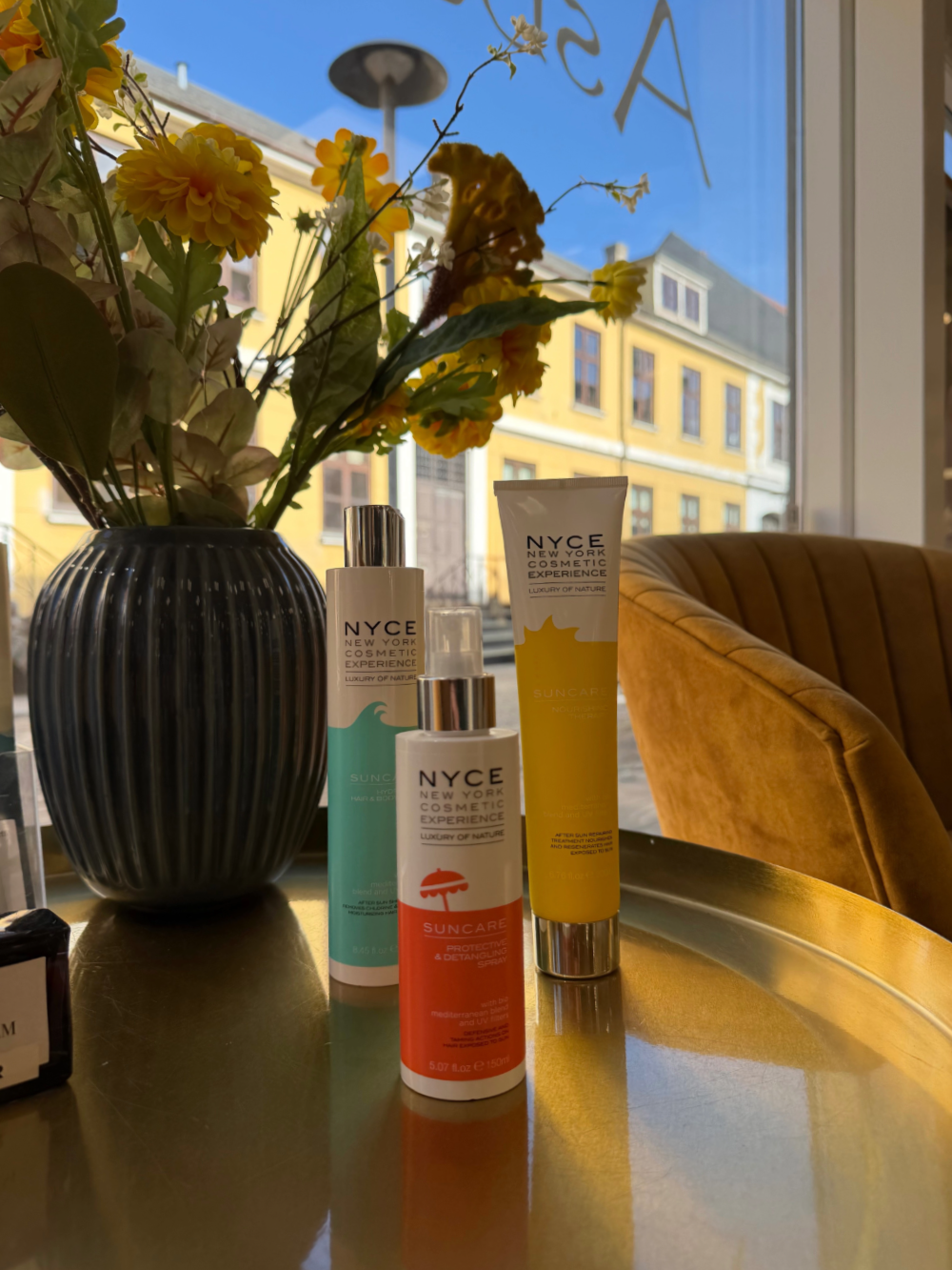 Tre hudplejeflasker på et solbeskinnet cafébord ved siden af ​​en vase med gule blomster.