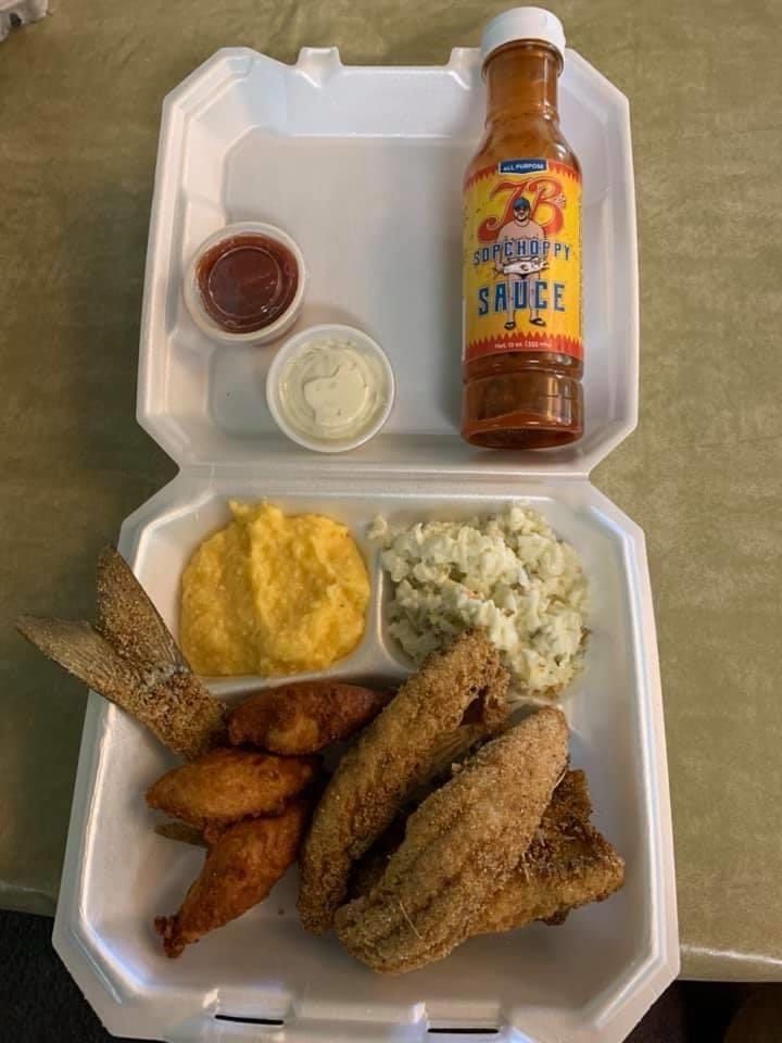 Takeout meal in a white clamshell container with fried fish, sides, sauces, and a bottle of hot sauce