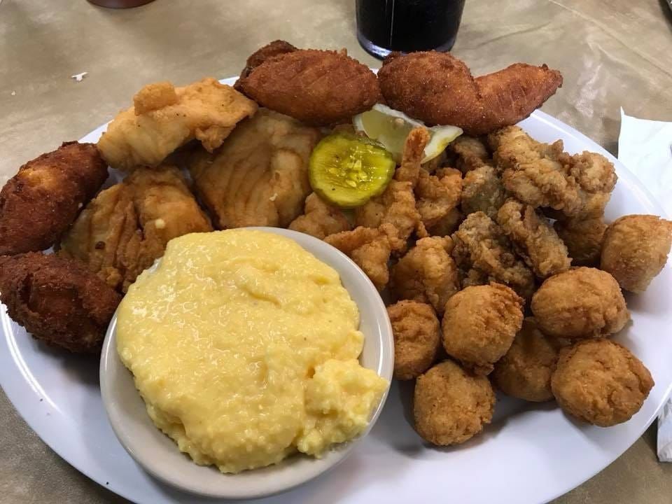 Plate of fried seafood and sides, including hush puppies, coleslaw, and grits, with pickles on top.