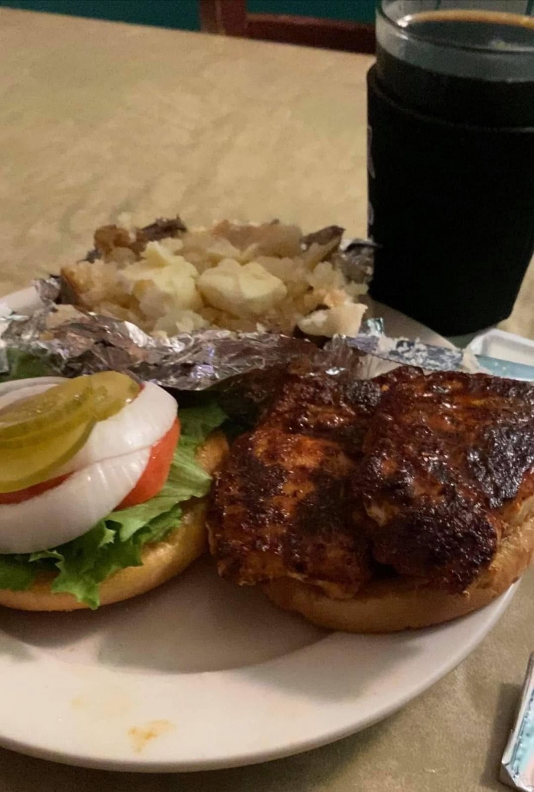 Grilled chicken, foil-wrapped baked potato, and open-faced sandwich with pickles on a plate beside a dark drink.