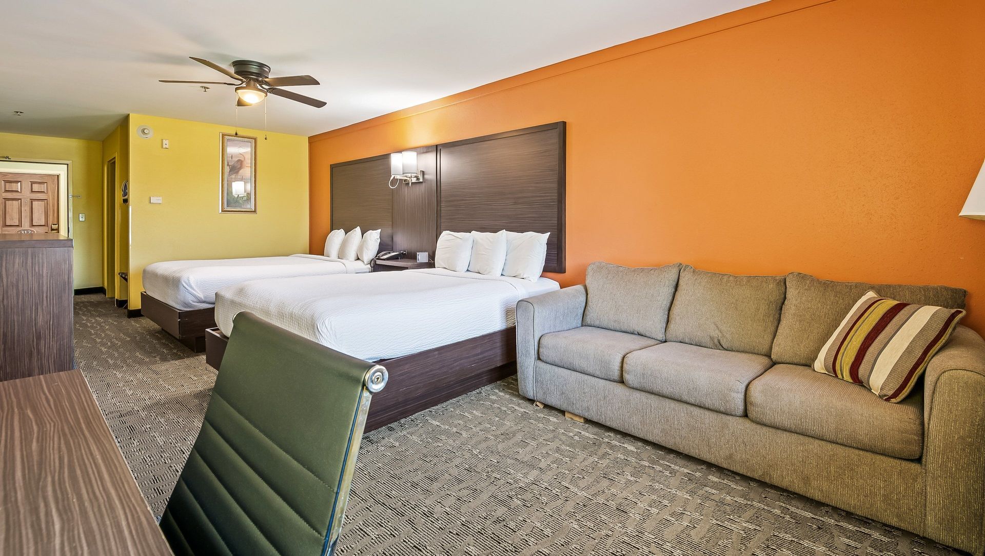 Hotel room with two beds, gray sofa, orange accent wall, and yellow hallway in the background