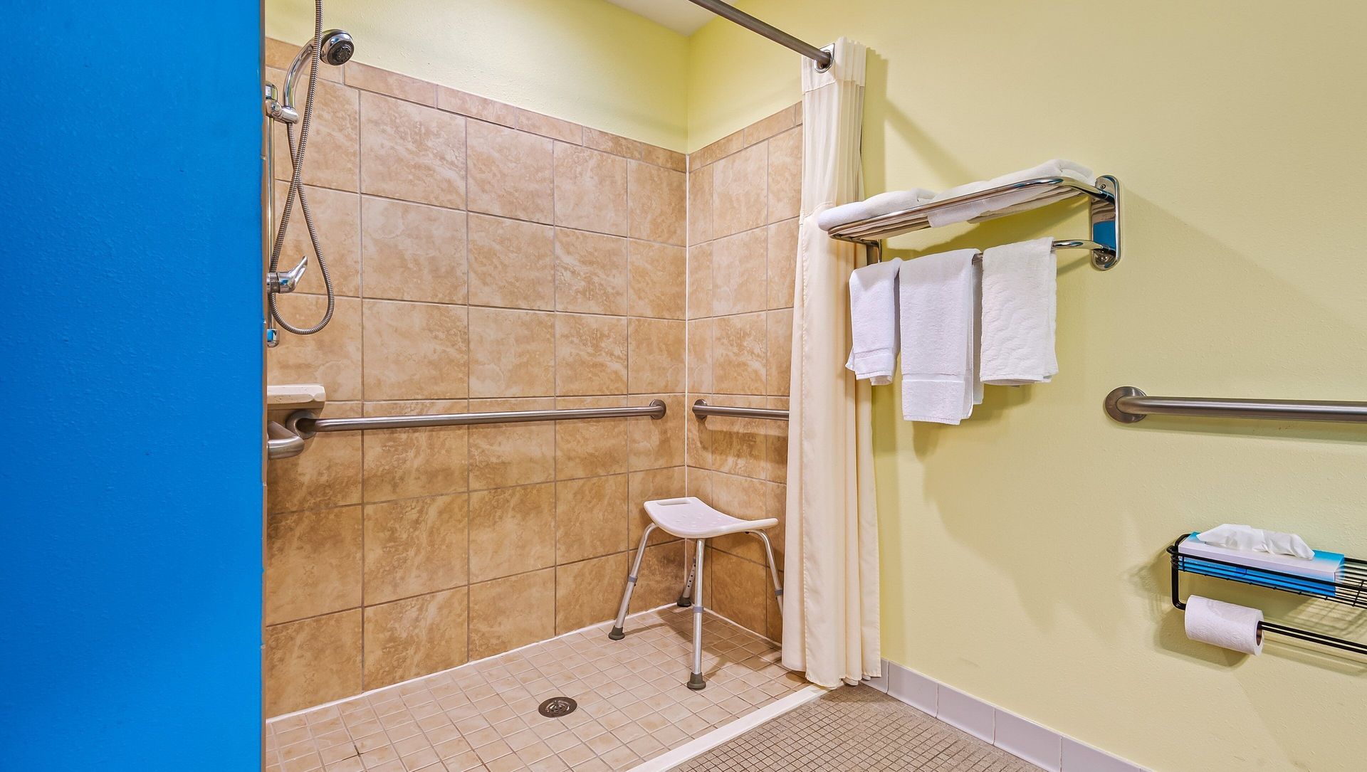 Accessible shower with tan tile walls, grab bars, fold-down seat, and a shower curtain