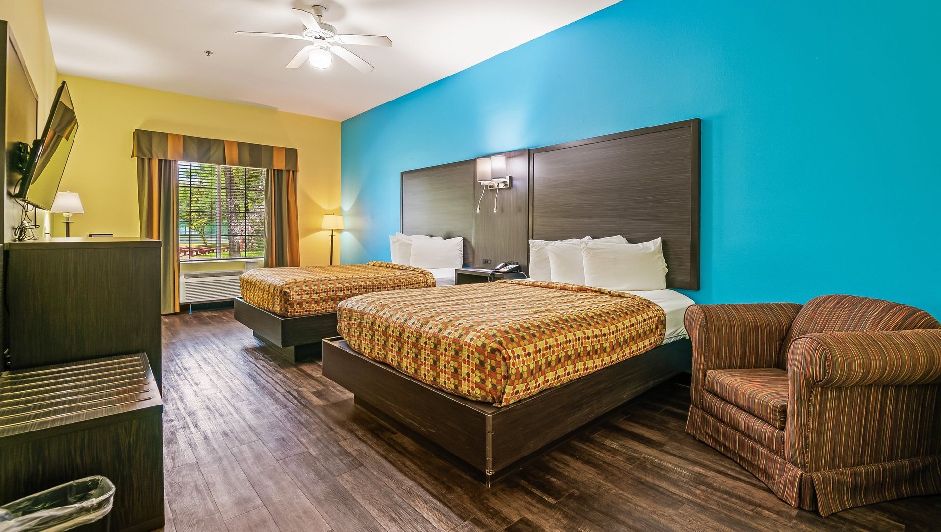 Bright hotel room with two beds, turquoise walls, dark wood floors, and a seating chair by the window