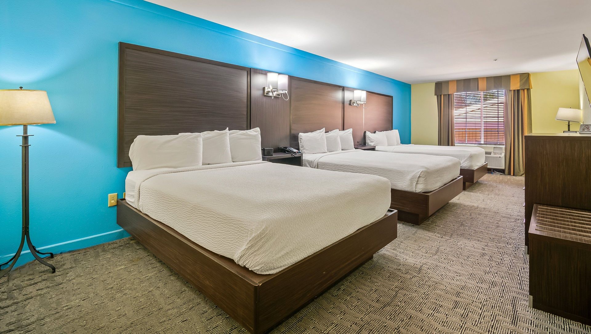 Hotel room with two queen beds, turquoise accent wall, floor lamp, and patterned carpet