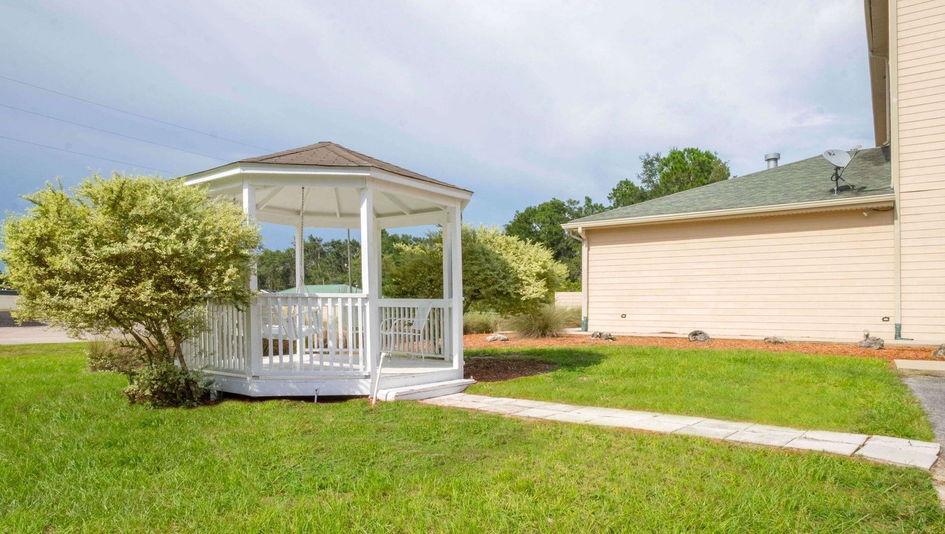 White gazebo in a green backyard beside a tan house, with a paved path and blue sky.