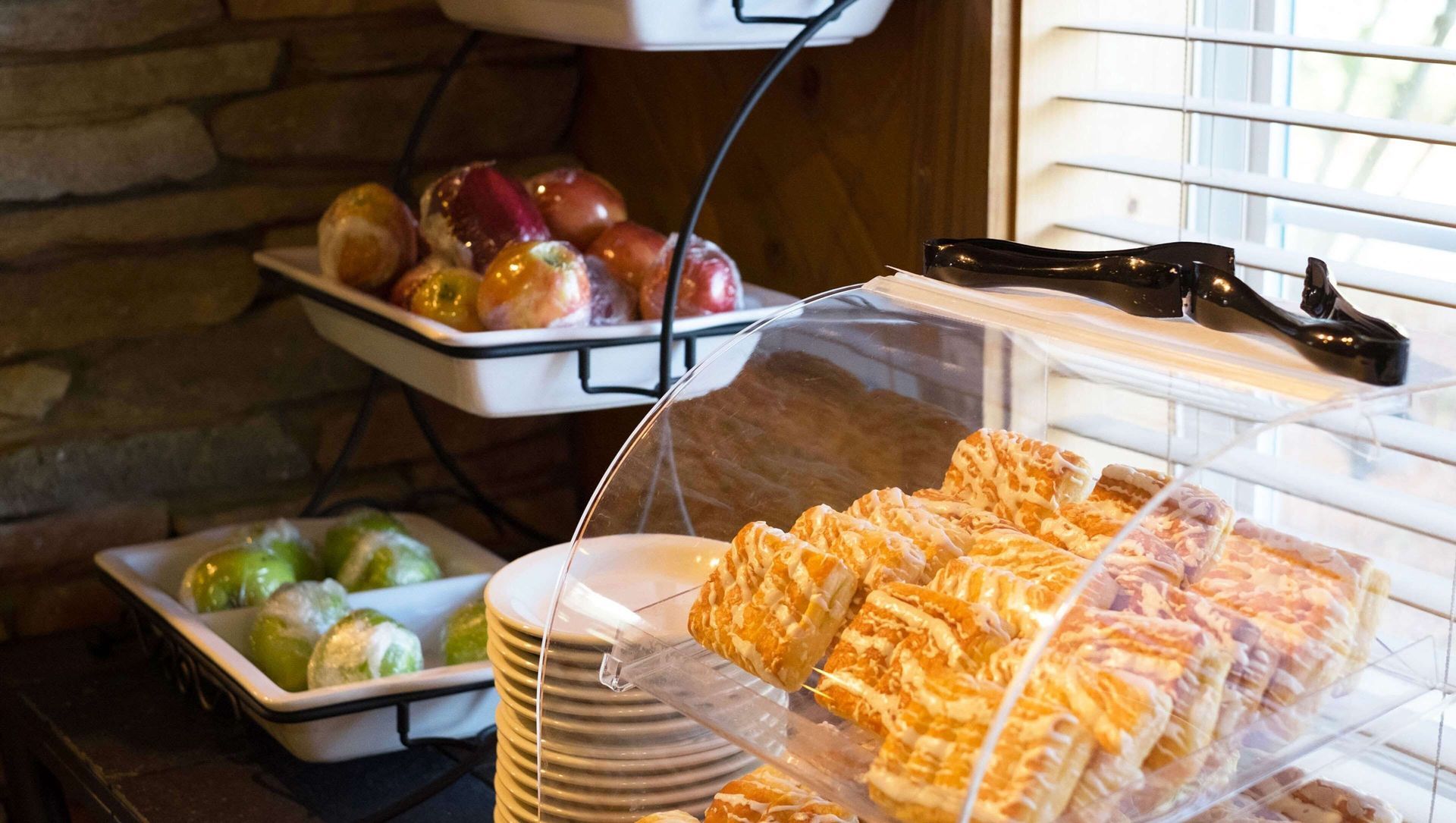 Bakery display case with pastries, stacked plates, and fruit in trays under a lighted shelf