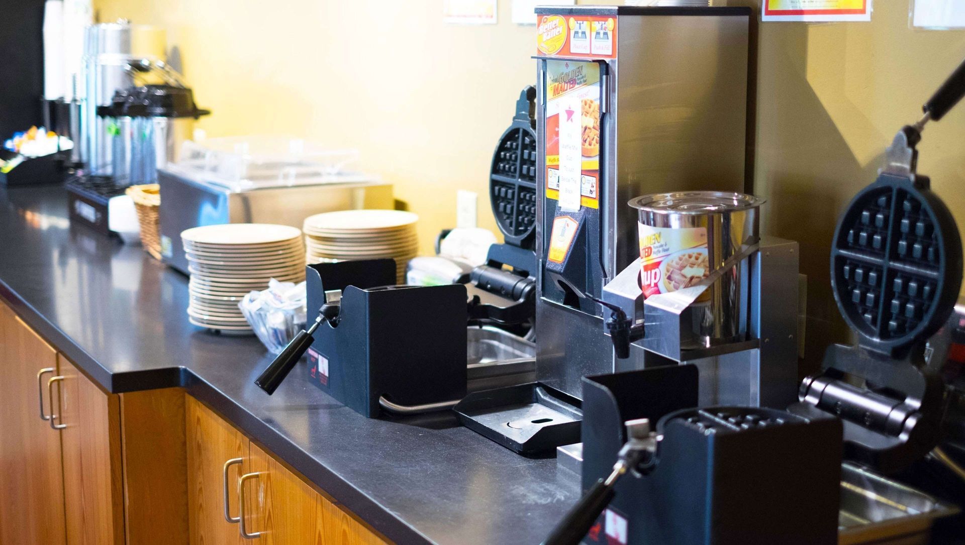 Counter with a coffee machine, cups, stacks of plates, and condiment stations in a cafeteria.