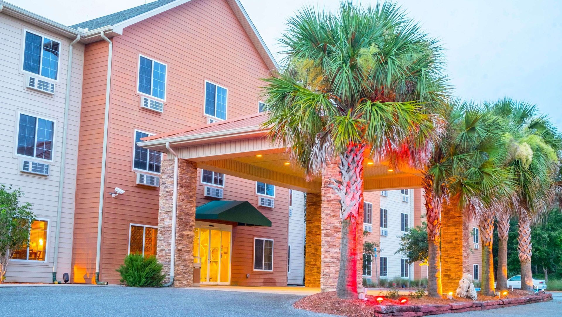 Brick hotel entrance with lit canopy, palm trees, and landscaped sidewalk at dusk