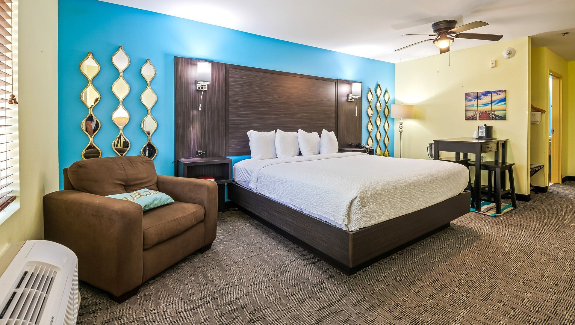 Hotel room with king bed, teal accent wall, armchair, desk, and window blinds