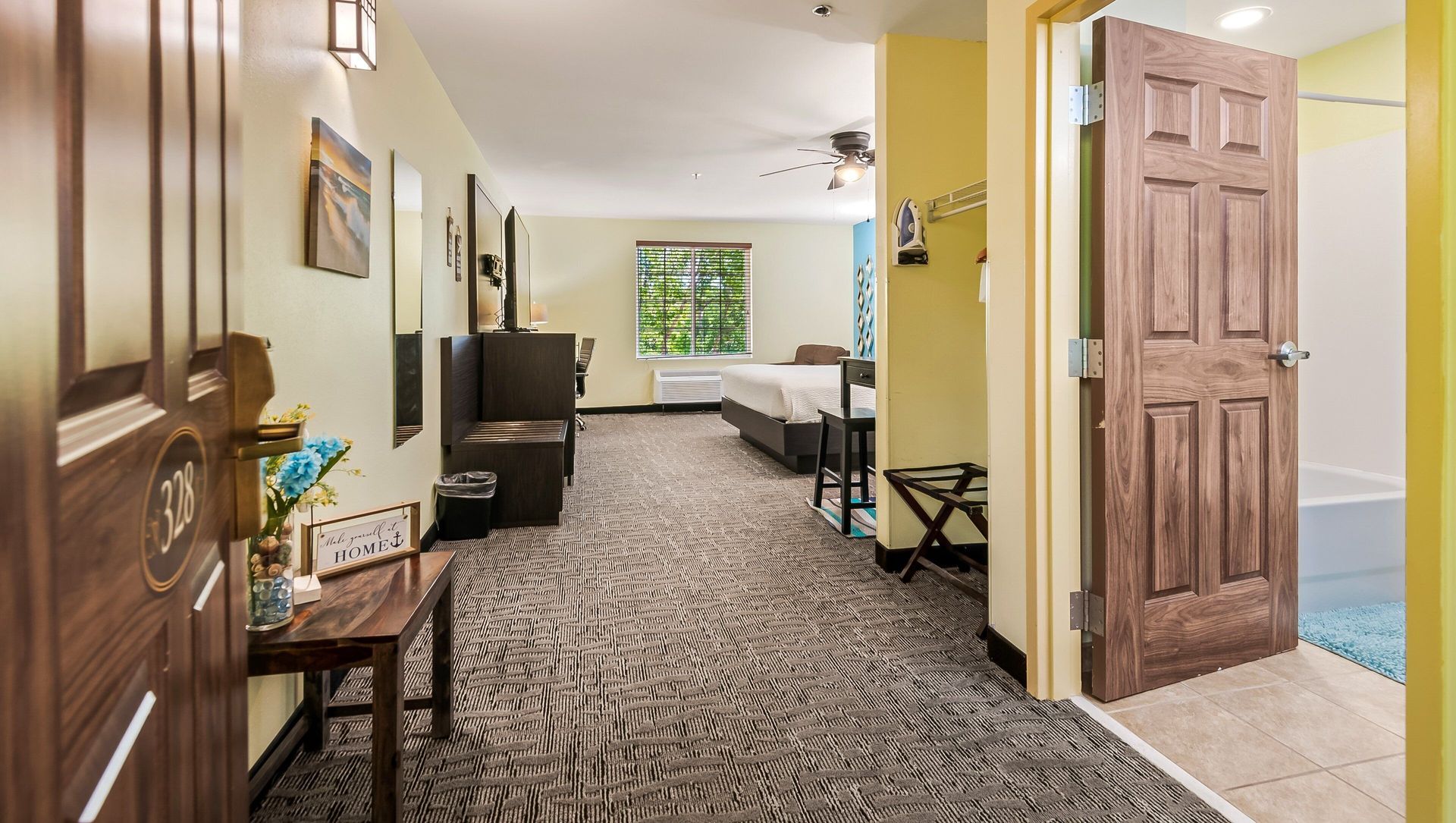 Hallway in a hotel room with patterned carpet, seating area, and open bathroom door