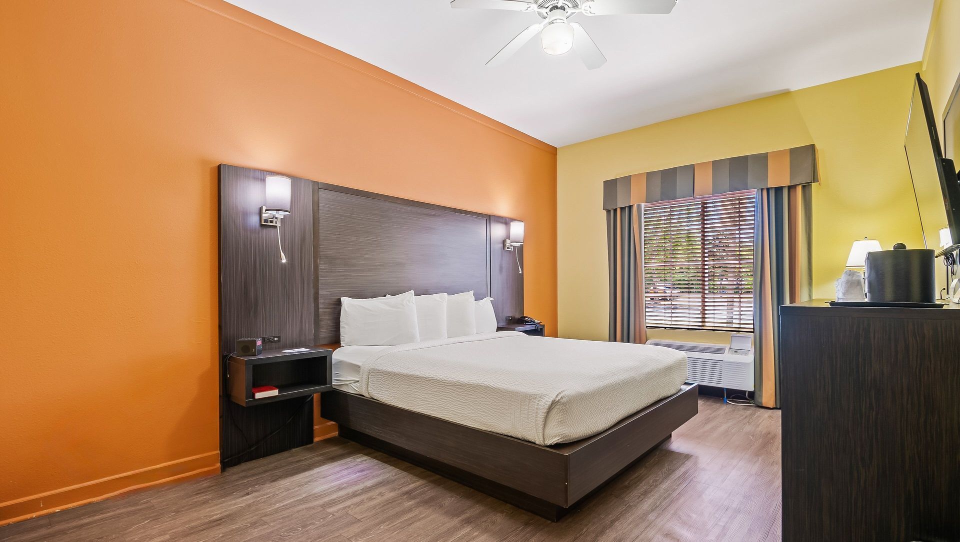 Modern hotel room with orange walls, a king bed, dark wood furniture, and a window with striped curtains