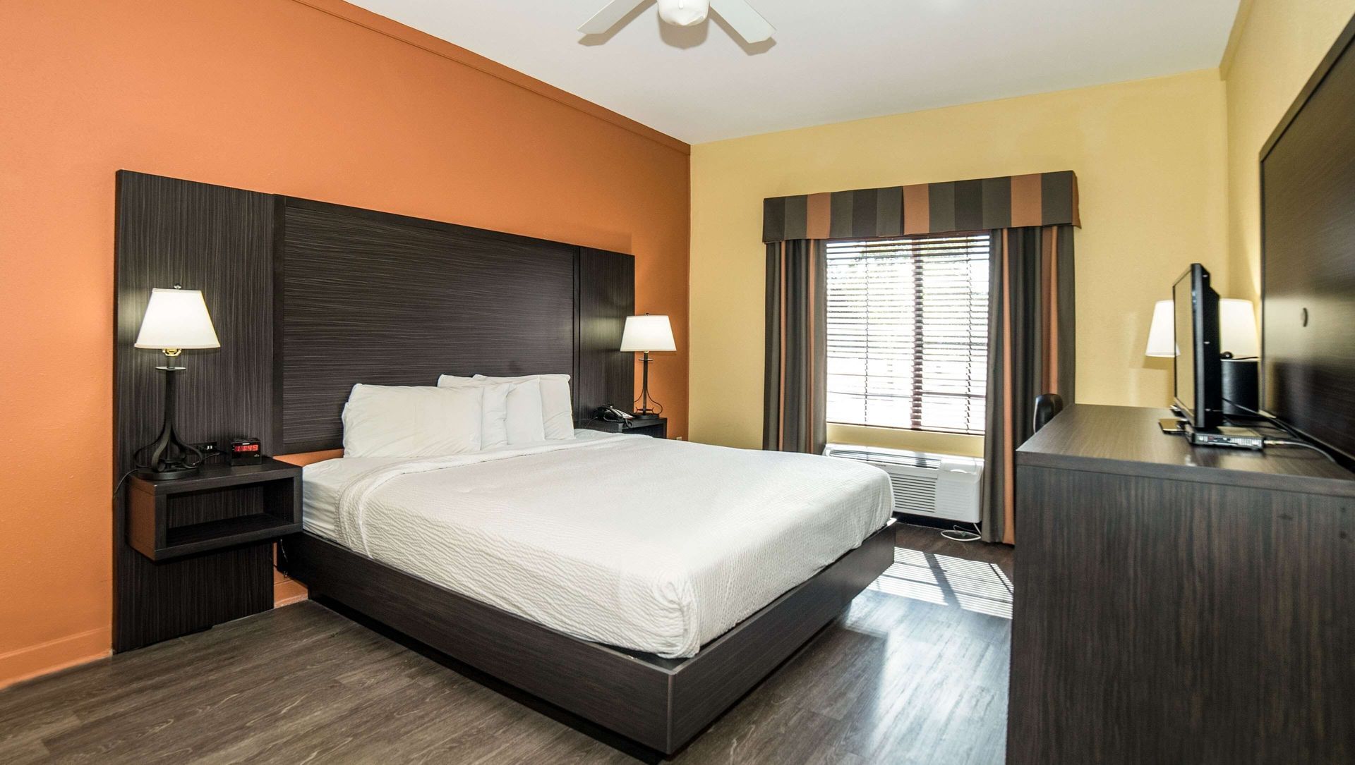 Hotel room with a neatly made bed, orange and cream walls, and a desk by the window