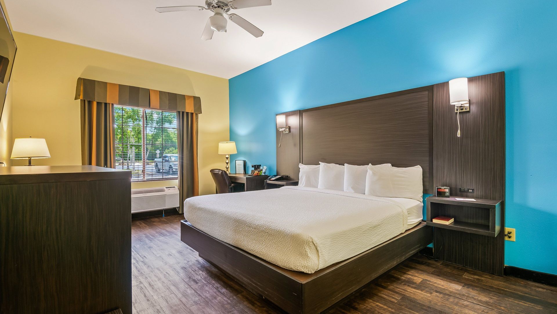 Hotel room with king bed, teal accent wall, wood flooring, and a window seat by a dresser.