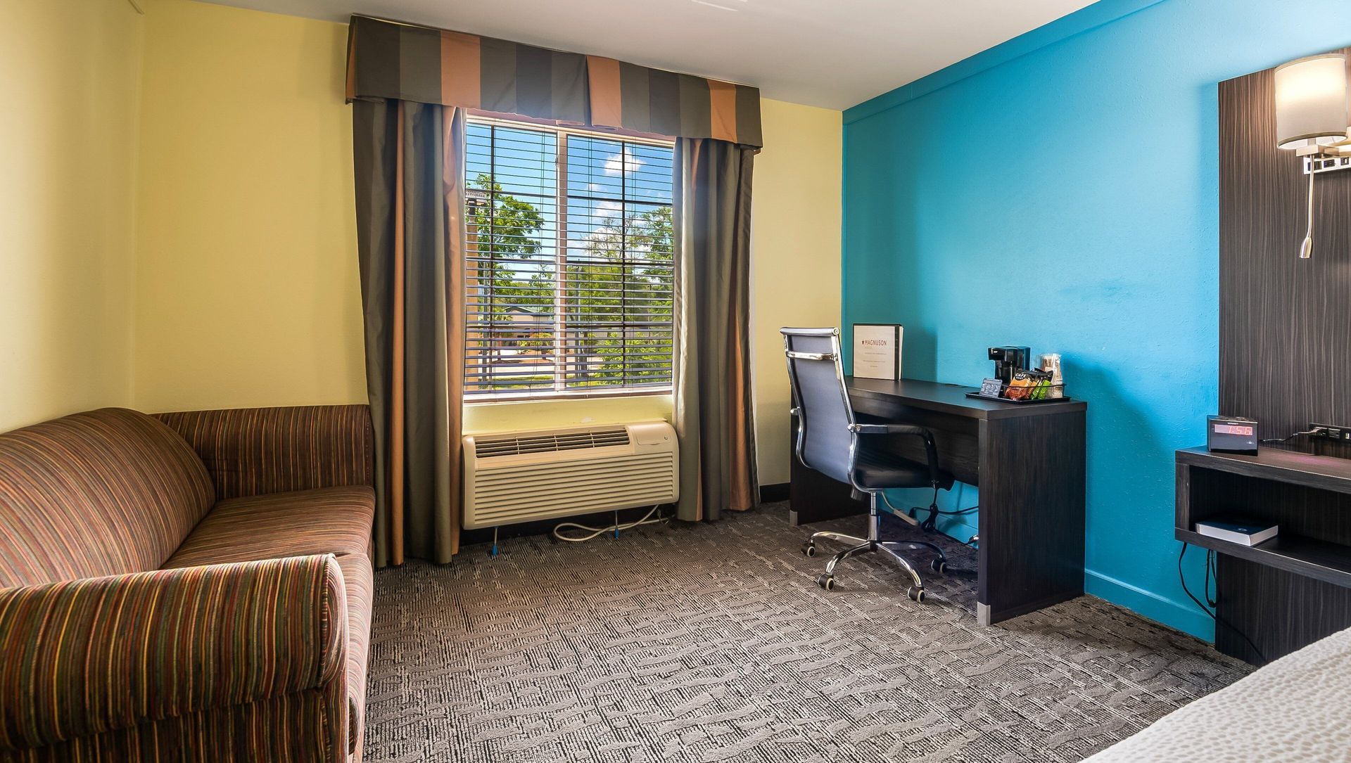 Hotel room with blue accent wall, desk and chair, sofa, patterned carpet, and window with curtains