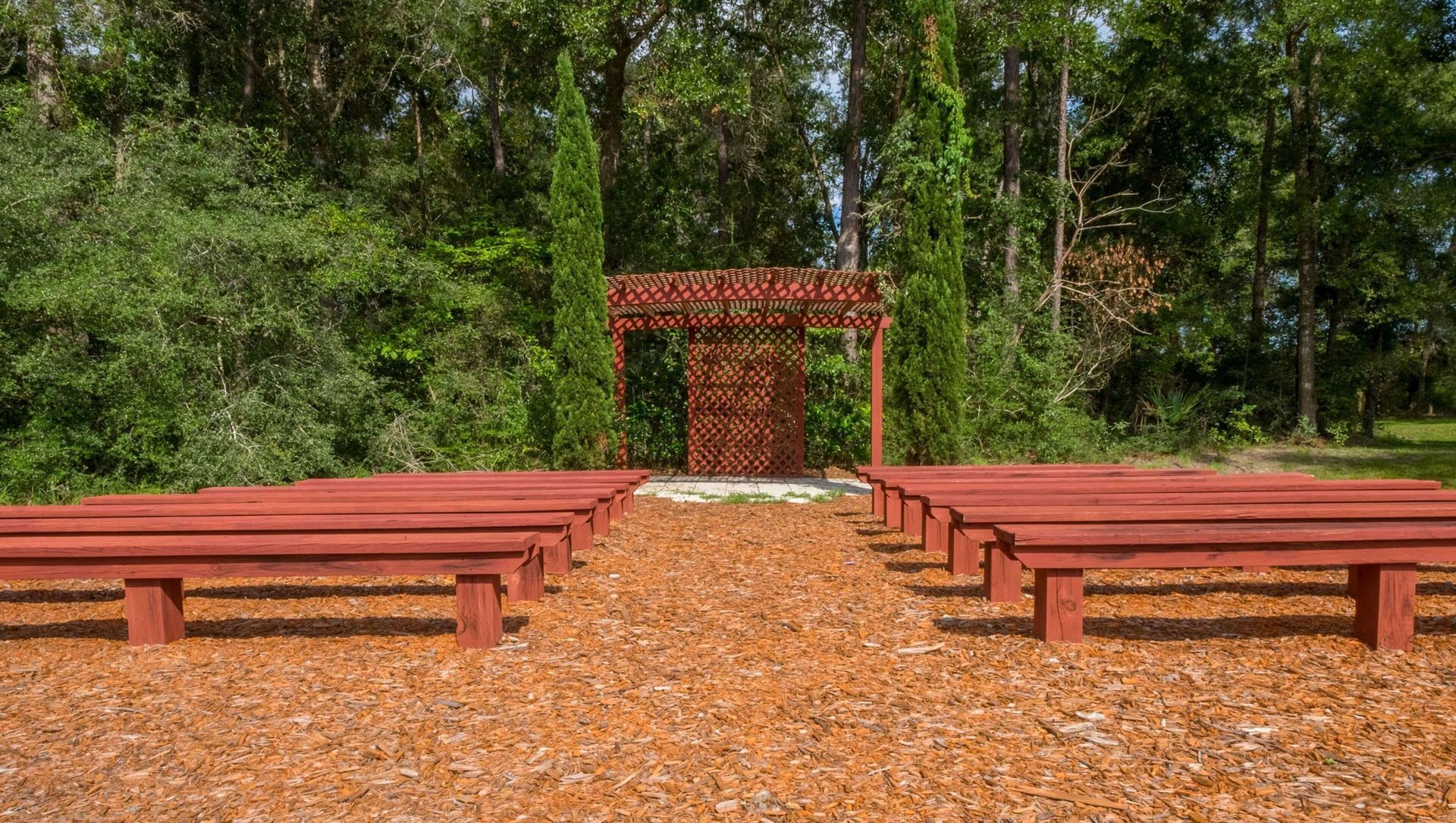 Woodland outdoor amphitheater with red benches, central aisle, and small wooden arch stage