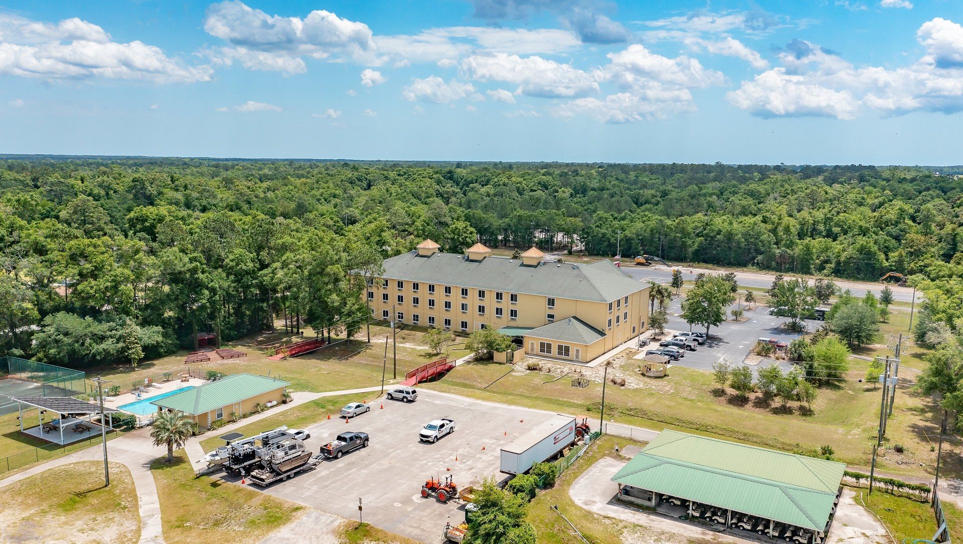 Aerial view of a beige building, parking lot, and pool surrounded by dense green trees.