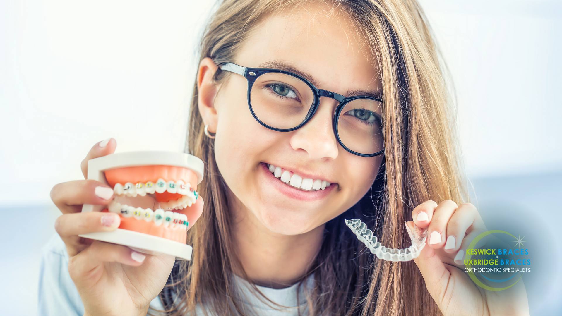 A young girl wearing glasses is holding a model of teeth and braces.