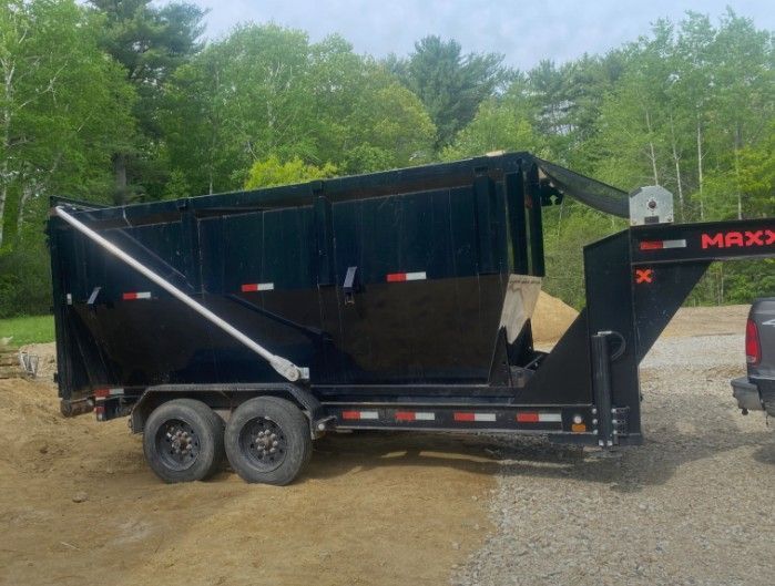 Black dump trailer with tarp cover and dual axles on a gravel lot, with trees in the background