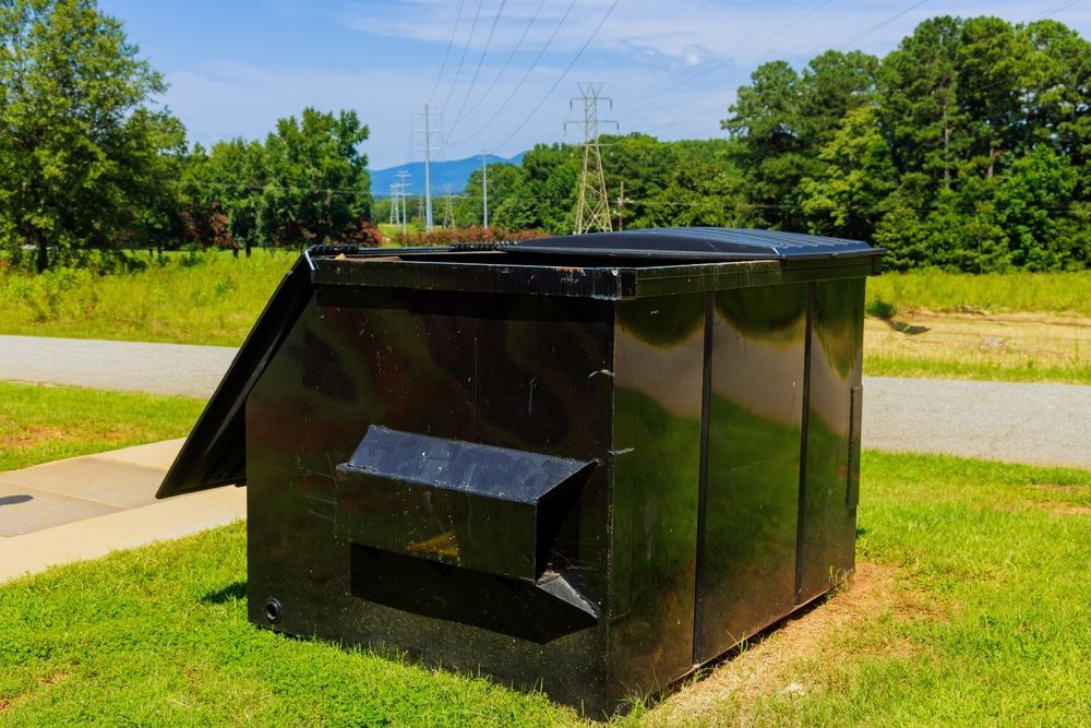 Black metal dumpster on grass beside a road, with trees and hills in the background.