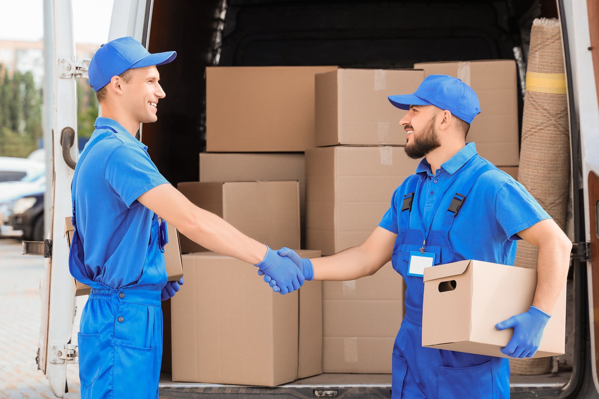 Two uniformed movers in blue shake hands beside stacked boxes in an open moving truck.