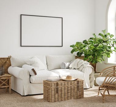 Bright living room with white sofa, wicker coffee table, potted plant, and empty wall frame