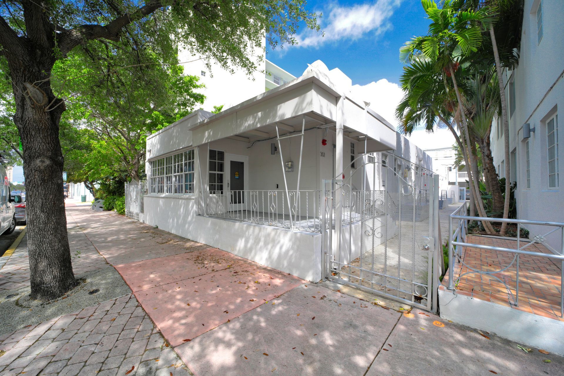 White house with a porch and gated walkway on a tree-lined street under a blue sky.