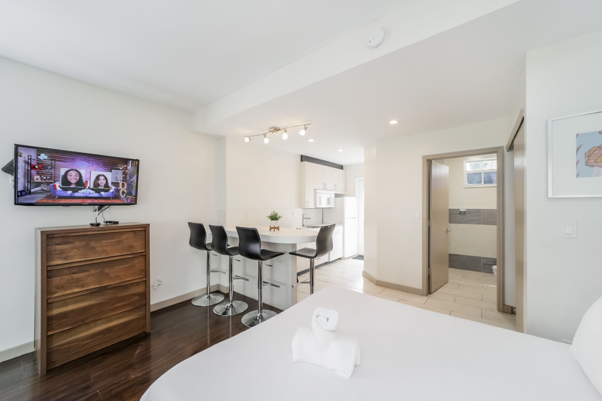 Bright bedroom with wall-mounted TV, bar stools by a table, and an open bathroom doorway.