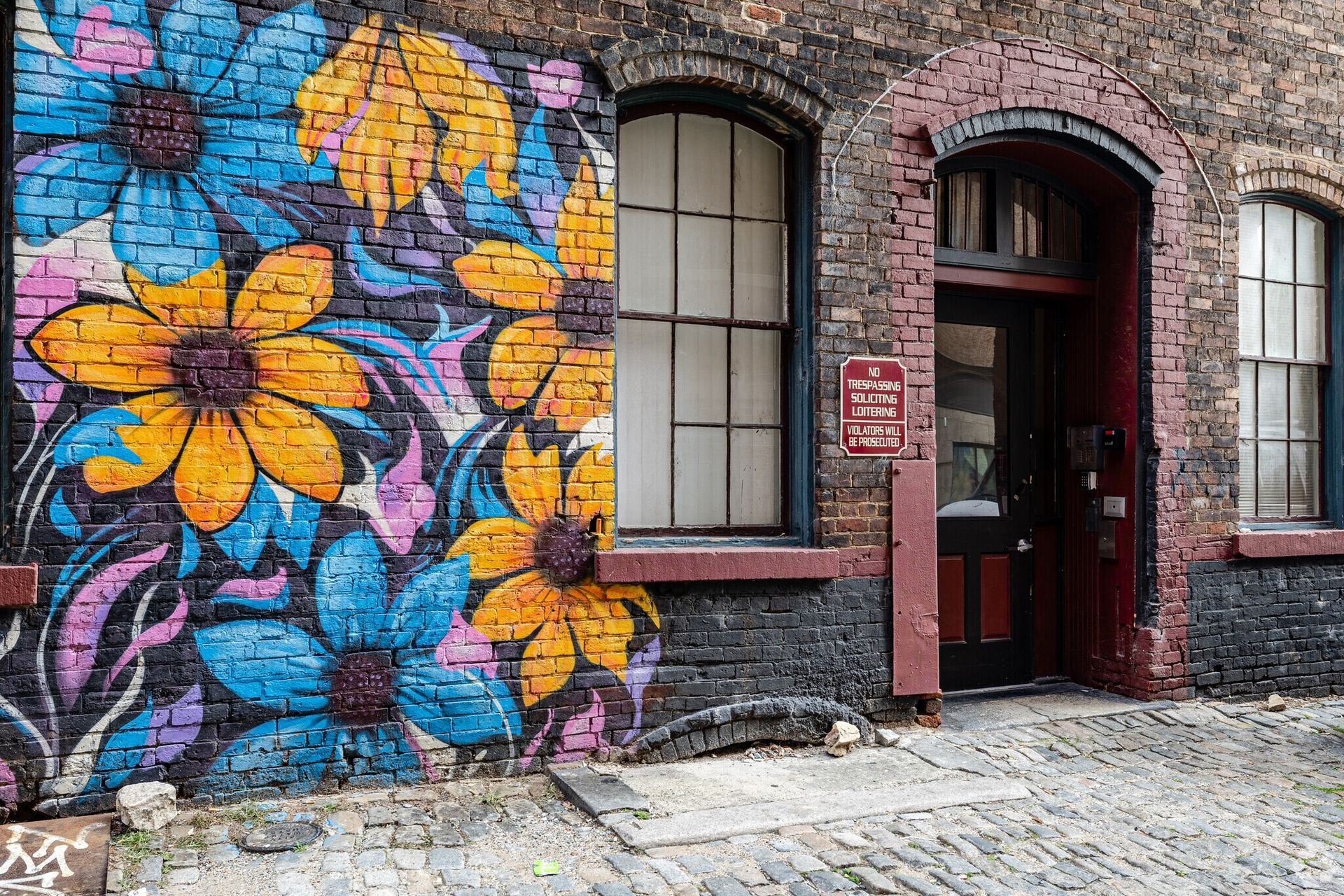 Brick building exterior with floral mural, window, doorway, and cobblestone path.