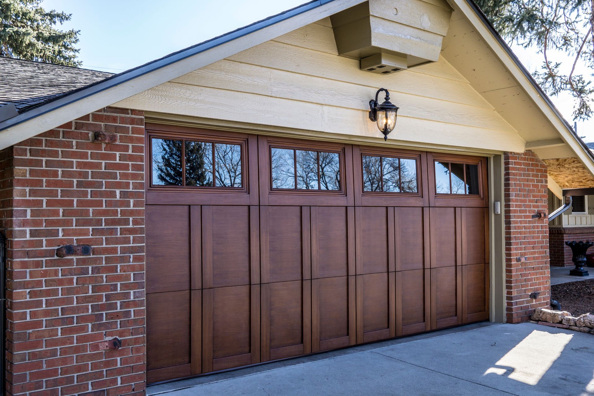 Brown wooden garage door on a brick house with a beige gable roof and wall light