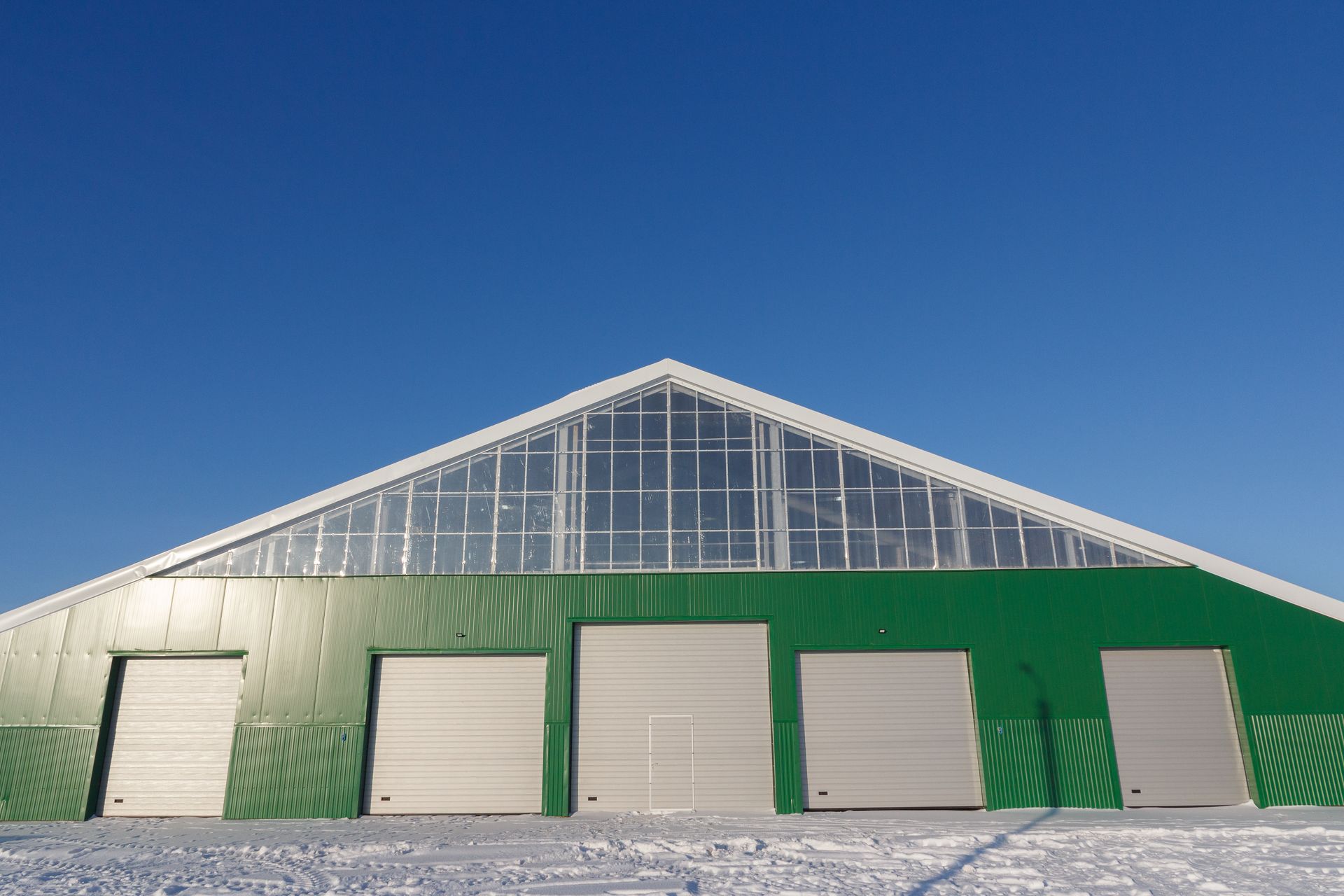 Green barn with five garage doors and a snow-covered roof under a clear blue sky