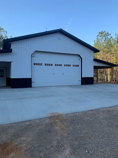 White metal garage with a large door and concrete driveway, set against trees under a clear blue sky