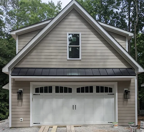 Tan house with white garage doors, dark metal awning, and a gabled roof among trees