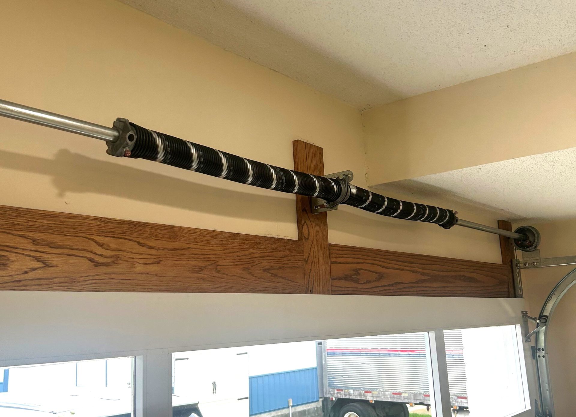 Garage door torsion spring wrapped in black coils on a metal rod above a wooden beam.