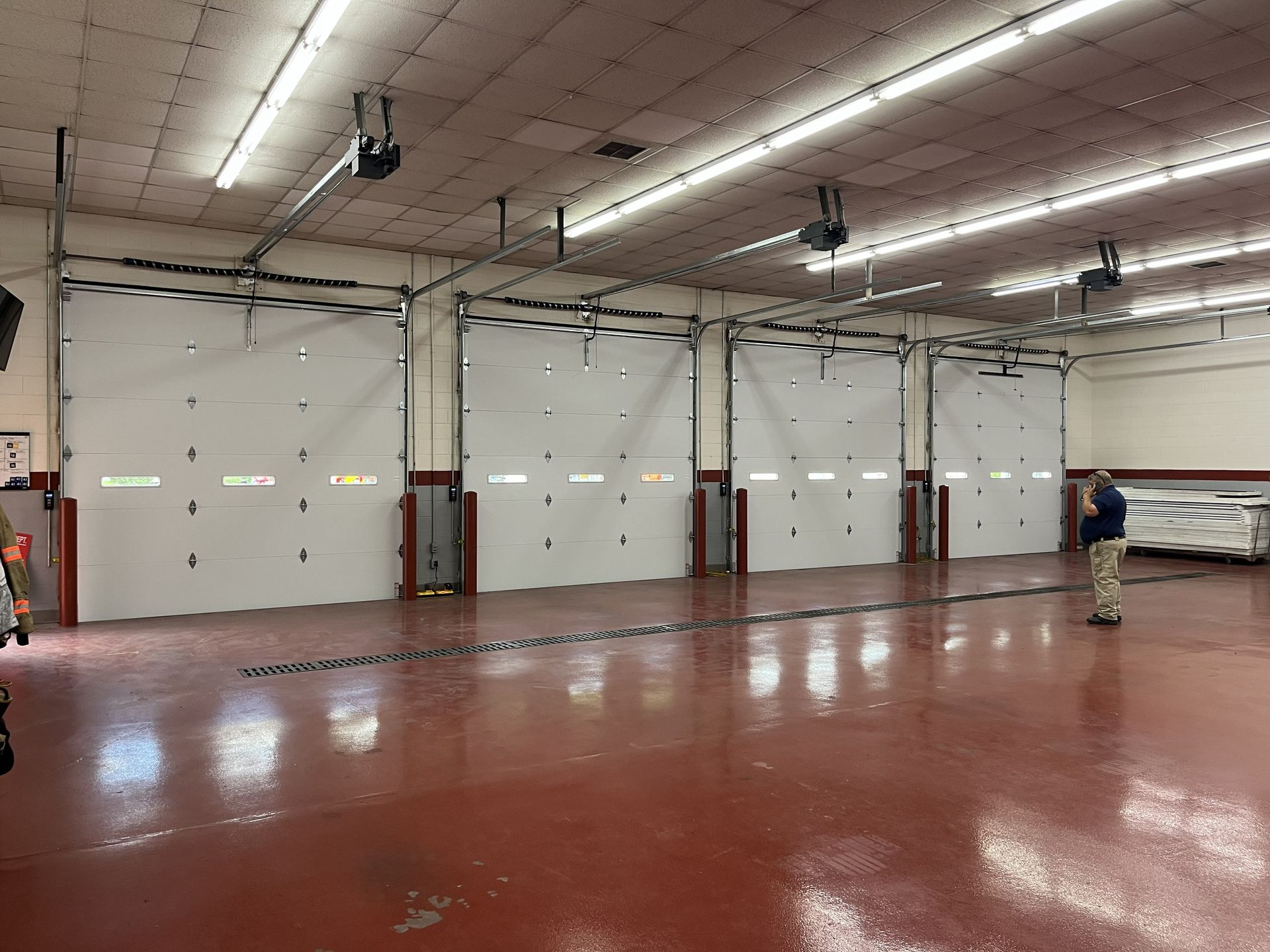 Large empty warehouse with a red floor, white roll-up doors, and a person standing near the right wall Large empty warehouse with a red floor, white roll-up doors, and a person standing near the right wall